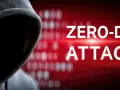 Sylwetka hakera w kapturze na tle czerwonego ekranu z napisem "ZERO-DAY ATTACK" i rozmytym kodem binarnym.