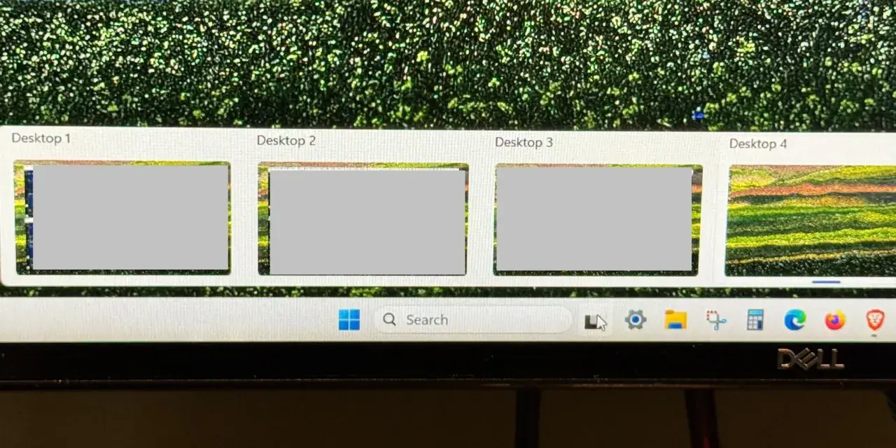 Widok czterech pulpitów Windows 10, z czego trzy są szare, a czwarty pokazuje kolorowy krajobraz. Pasek zadań z wyszukiwarką i ikonami.