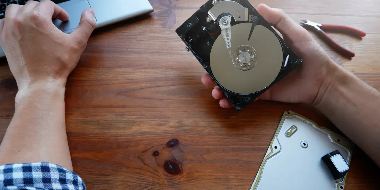 Ręce majstrują przy dysku HDD, obok laptop i kombinerki. Dowiedz się, jak przyspieszyć dysk HDD.