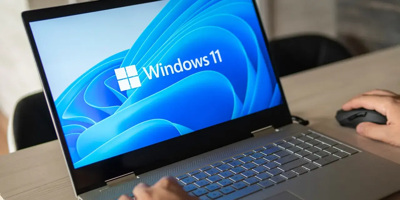 Pierwsze uruchomienie komputera z systemem Windows 11. Dłoń użytkownika przesuwa myszkę, a ekran laptopa wyświetla logo systemu.