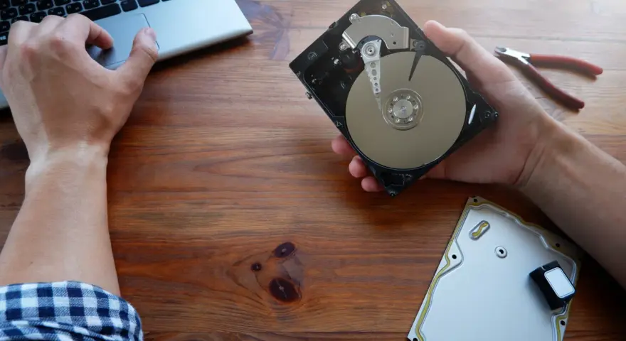 Ręce majstrują przy dysku HDD, obok laptop i kombinerki. Dowiedz się, jak przyspieszyć dysk HDD.