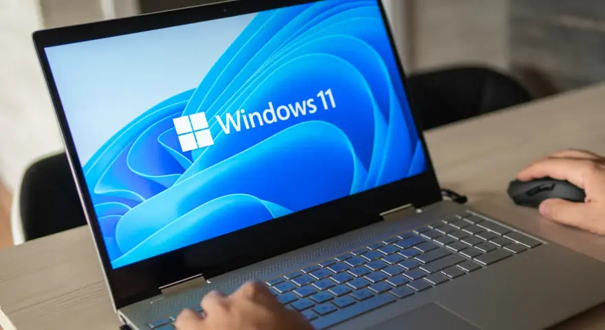 Pierwsze uruchomienie komputera z systemem Windows 11. Dłoń użytkownika przesuwa myszkę, a ekran laptopa wyświetla logo systemu.