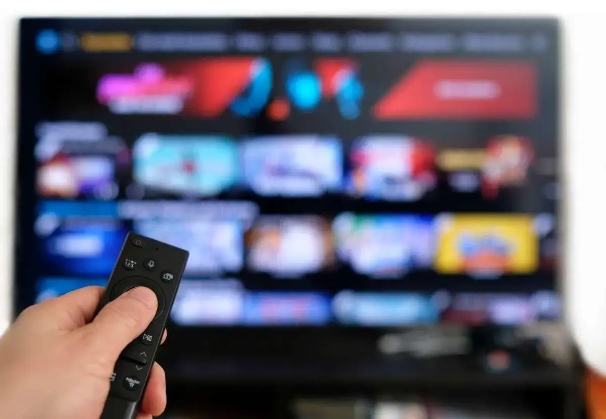 Dłoń trzymająca pilot do telewizora, na ekranie widoczne menu z filmami i serialami, jakby z vault 7.