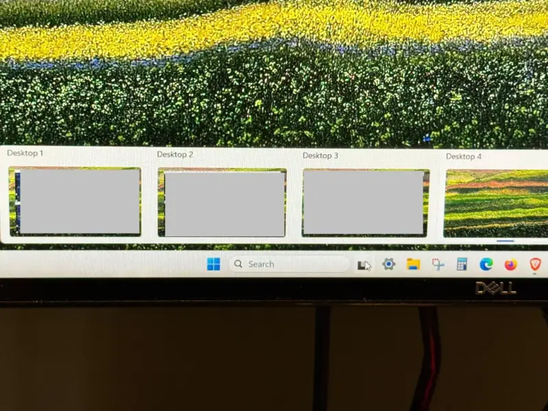Widok czterech pulpitów Windows 10, z czego trzy są szare, a czwarty pokazuje kolorowy krajobraz. Pasek zadań z wyszukiwarką i ikonami.