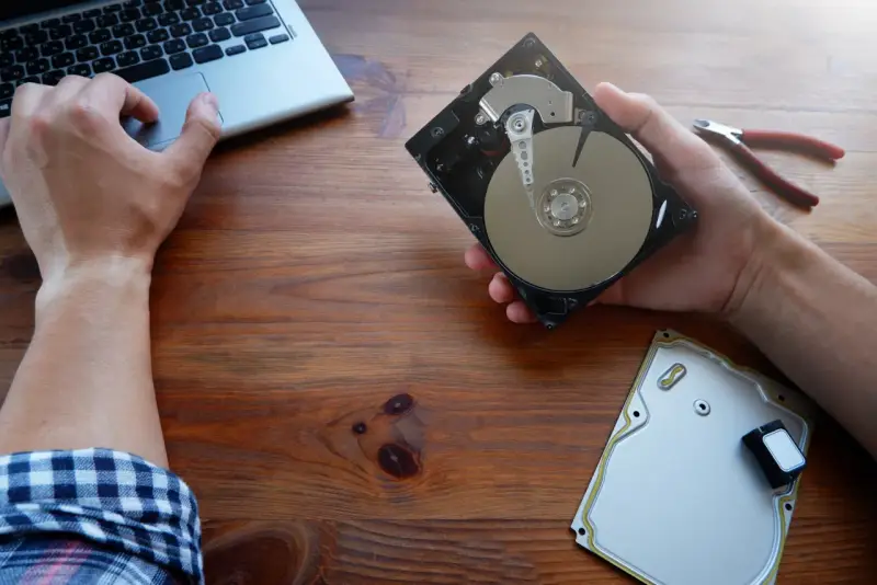 Ręce majstrują przy dysku HDD, obok laptop i kombinerki. Dowiedz się, jak przyspieszyć dysk HDD.